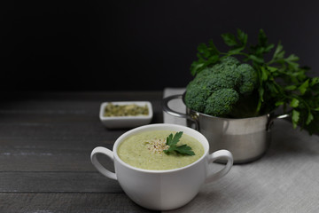 Vegetarian cream of broccoli, potatoes, onions, garlic, sesame and olive oil, and coconut milk soup served in a bowl with seeds on a dark brown plank background using gray textile