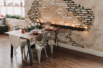 Wooden Dinner Table with Plate and Metallic Chairs. Decorative Tree Branch, Glasses and Champagne Bucket on Desk. Glowing Garland and Kitchen Ware on Windowsill. Old Brick Wall on Background