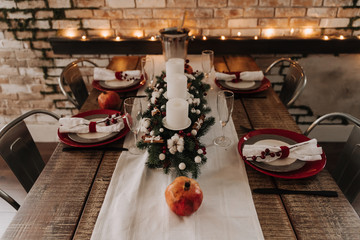 Xmas Wooden Dinner Table Decorated Candlestick. Decorative Christmas Tree Branch Ornamented Berries and White Balls, Glasses, Plates and Fruit. Grunge Brick Wall and Sparkle Garland on Background