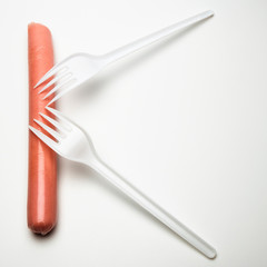A sausage and two forks on a white background look like the letter K . The concept of fast food, hunger or lack of food. Environmental issue and ban on plastic dishes, poor student or senior citizen.