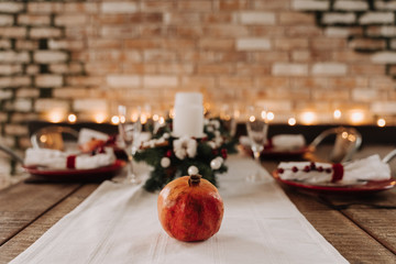 Pomegranate on Holiday Christmas Dinner Table. Ripe Fruit on White Tablecloth. Xmas Tree Branch, Champagne Glasses, Plates with Cutlery. Antique Brick Wall with Moss and Lighting Garland on Background