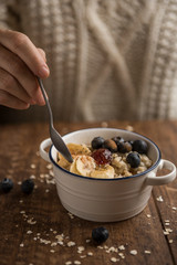 Healthy breakfast on wooden table, woman's hand and porridge with oat flakes, banana, blueberries, chia, cinnamon, maple syrup and strawberry jam. Vertical with copyspace
