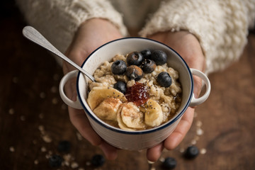 Woman hands offering a vegan porridge with oat flakes, banana, blueberries, chia, cinnamon, maple syrup and strawberry jam. Horizontal. Top view. - obrazy, fototapety, plakaty