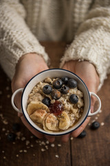 Woman hands holding a vegan breakfast, porridge with oat flakes, banana, blueberries, chia, cinnamon, maple syrup and strawberry jam. Rustic wooden table in the background. Vertical top view.