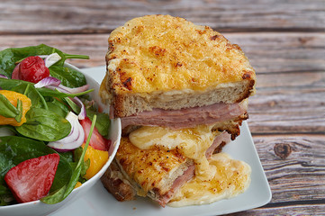 Sandwich with Parmesan and Gruyere cheese, ham, béchamel sauce and artisan bread accompanied by a salad on a wooden background.  The Croque Monsieur.