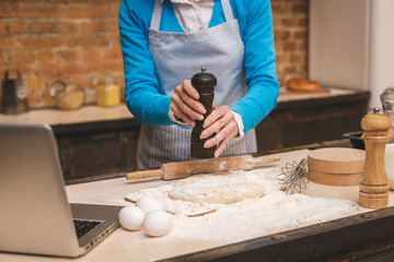 Attractive senior aged woman is cooking on kitchen. Grandmother making tasty baking. Using laptop. Close-up food concept.