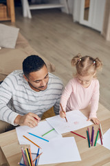 Naklejka premium High angle portrait of contemporary young dad drawing pictures together with cute little daughter at home, copy space
