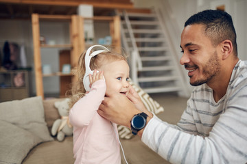 Side view portrait of contemporary dad giving big headphones to cute toddler while playing with kid at home, copy space