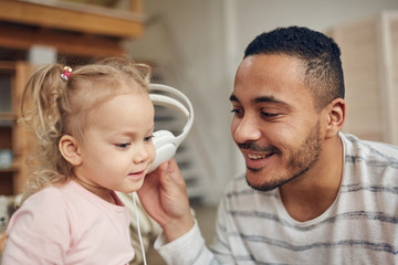 Portrait of contemporary mixed-race dad listening to music with cute little daughter via big headphones, copy space