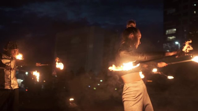 Confident female asian dancer moves with flaming torches with group of fireshow artists. They performing synchronously choreography while the camera goes around them. Slow motion.