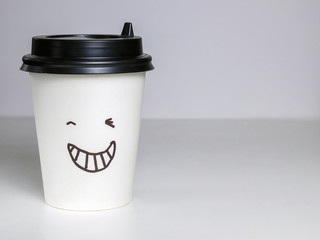 a glass of hot strong coffee or tea made of paper with a black lid and a winking face stands on a gray background, emotion on his face, a smile in his mouth. joyful, happy emotions