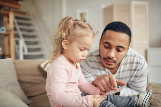 Portrait of caring young father looking at pictures while playing with cute little daughter at home, copy space