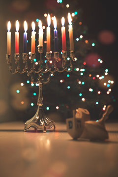 Hanukkah traditional chandelier (menorah) burning candles and spinning top toys (dreidels) on the background of Christmas tree with colorful lights.