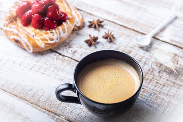Food on a wooden table made of whitish boards, a black mug of coffee, a puff of raspberries and icing, and a star anise, and a white ceramic spoon