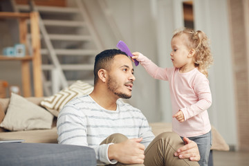 Fototapeta premium Portrait of cute little girl brushing hair of mixed-race dad while laying beauty salon, copy space
