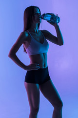Fototapeta na wymiar Young sporty muscular woman drinking water, isolated against purple light background