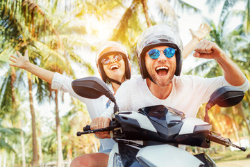 Fototapeta Happy smiling couple travelers riding motorbike scooter in safety helmets during tropical vacation under palm trees obraz