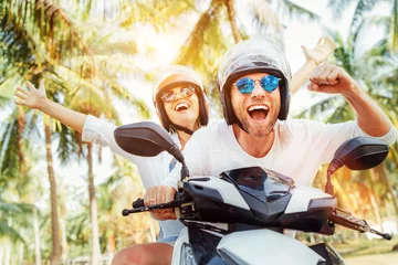 Poster Happy smiling couple travelers riding motorbike scooter in safety helmets during tropical vacation under palm trees © Soloviova Liudmyla