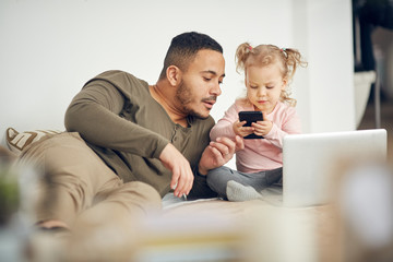 Portrait of cute little girl playing with father and using smartphone in cozy bedroom interior,...