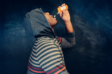 african american male pouring pills into mouth wearing hooded shirt against a dark background