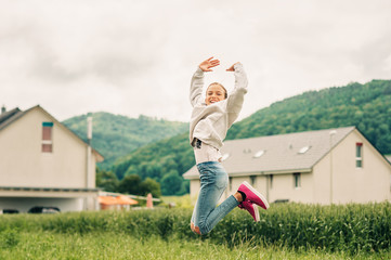Outdoor portrait of happy jumpink kid girl, wearing grey sweatshirt, denim jeans and pink shoes