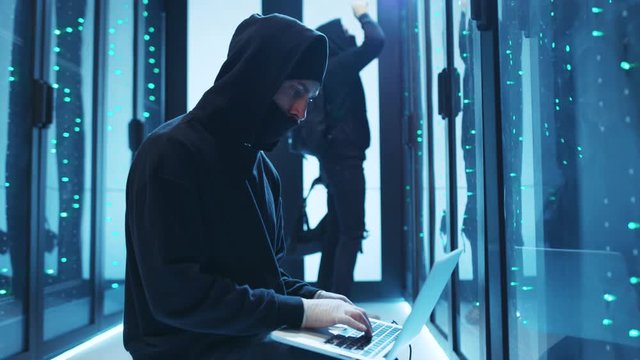Hooded hacker coding phishing attacks on laptop breaking into secure server cabinet. Team of professional spies stealing information from database at modern data center.