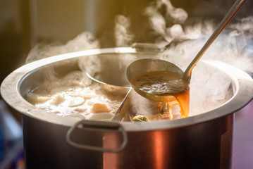 Cooking person in the restaurant is cooking while using the dipper in a large soup pot. And the water is boiling, there is smoke and hot spit.