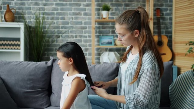 Caring mother attractive young woman is is brushing daughter's hair taking care of beautiful child at home sitting on couch together. Beauty, childhood and house concept.