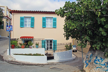 The front of a beautiful typical home with aqua shutters and gated courtyard entrance on a rounded corner along a winding, hilly, narrow, Old World street in Anafiotika, Athens, Greece.