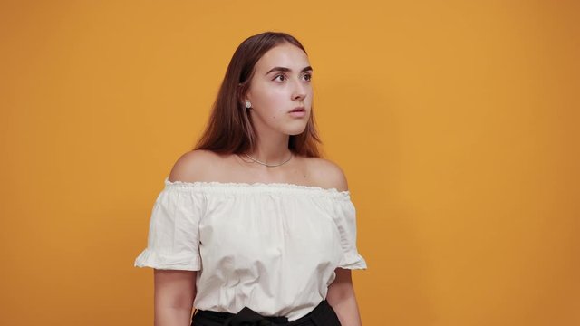 Disappointed young woman doing pointing into distance, looking serious isolated on orange background in studio in casual white shirt. People sincere emotions, lifestyle concept.