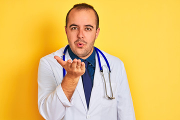 Young doctor man wearing coat and stethoscope standing over isolated yellow background looking at the camera blowing a kiss with hand on air being lovely and sexy. Love expression.