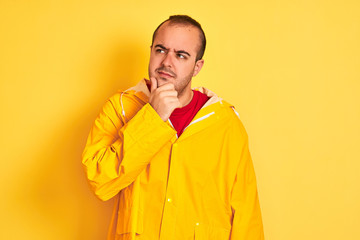 Young man wearing rain coat standing over isolated yellow background with hand on chin thinking about question, pensive expression. Smiling with thoughtful face. Doubt concept.