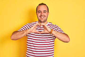 Young man wearing casual striped t-shirt standing over isolated yellow background smiling in love showing heart symbol and shape with hands. Romantic concept.