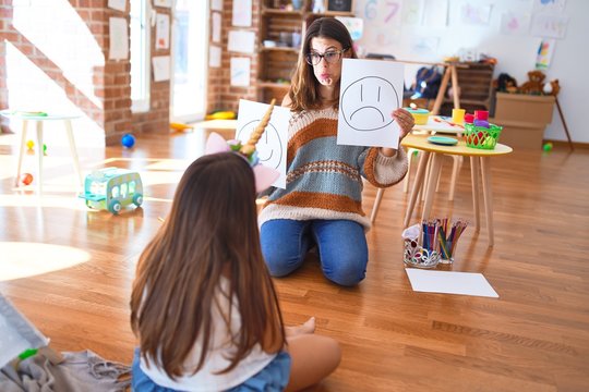 Beautiful pshychologist and toddler doing therapy with emotions emoji around lots of toys at kindergarten