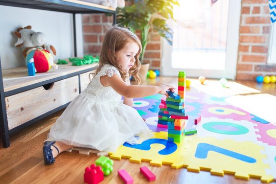Adorable blonde toddler playing with building blocks toy around lots of toys at kindergarten