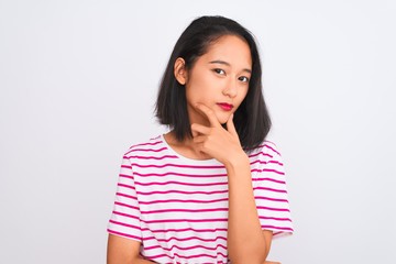 Young chinese woman wearing striped t-shirt standing over isolated white background looking confident at the camera with smile with crossed arms and hand raised on chin. Thinking positive.