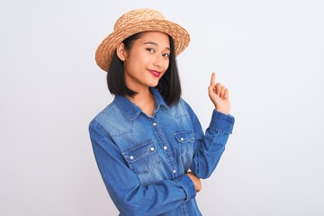 Young beautiful chinese woman wearing denim shirt and hat over isolated white background with a big smile on face, pointing with hand and finger to the side looking at the camera.