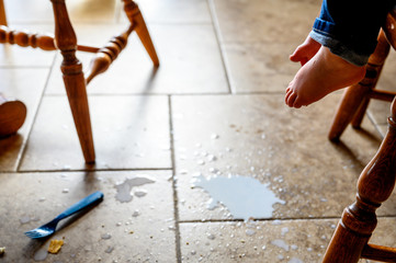 toddler foot suspended above spilled milk, crumbs, and a plastic fork on a brown tile floor 