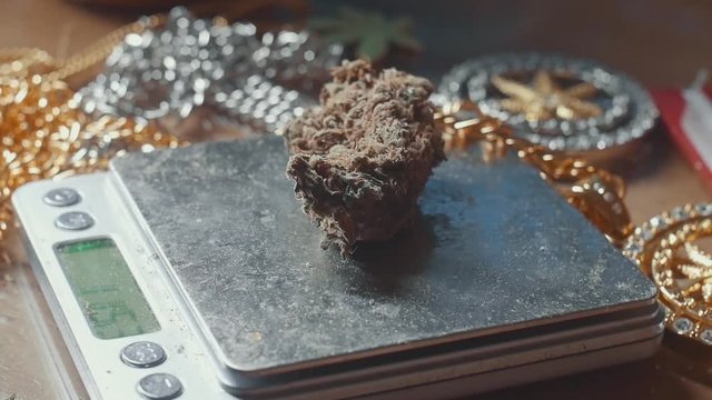 Handheld close-up of hand putting chunks of weed on a scale that's laying on a table covered by gold chains