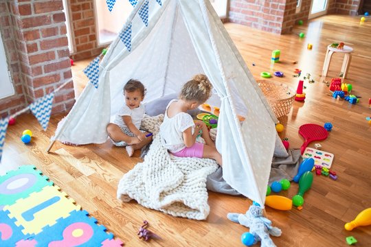 Beautiful toddlers playing inside tipi over blanket around lots of toys at kindergarten