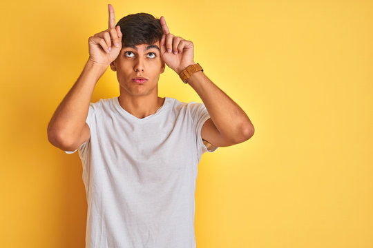 Young indian man wearing white t-shirt standing over isolated yellow background doing funny gesture with finger over head as bull horns