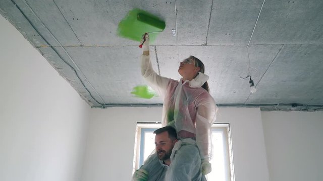 Dolly cam: young bearded man holds beautiful woman in working suit painting ceiling with green color in spacious room