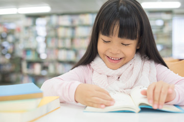 portrait of cute schoolgirl smiling while sitting with stack of books at table in library