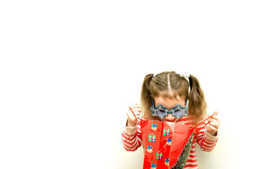 isolated little girl in red sweater and blue snowflake-like glasses looking into red gift bag on white background with copy space