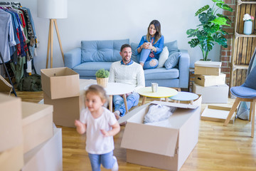 Beautiful family, parents sitting on the sofa drinking coffee looking his kid playing at new home around cardboard boxes