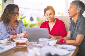 Meeting of middle age women having lunch and drinking coffee. Mature friends smiling happy using laptop at home on a sunny day