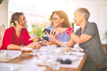 Meeting of middle age women having lunch and drinking coffee. Mature friends smiling happy using smartphone at home on a sunny day