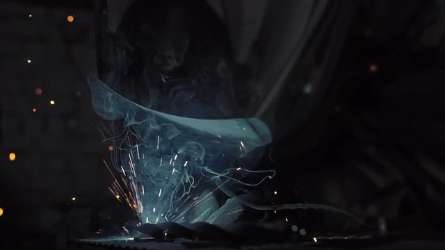 Forge workshop. Smithy. Worker in a welding hood helmet welds a part by electric welding. Sparks are reflected in the protective screen. Blacksmith makes iron product for manufacture. Slow motion.