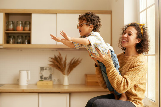 Portrait Of Cute Young Latin Woman In Sweater Sitting On Wndowsill Holding Her Two Year Old Son Who Is Reaching Out Hands As If Flying. Happy Mom And Child Playing In Cozy Kitchen Interior