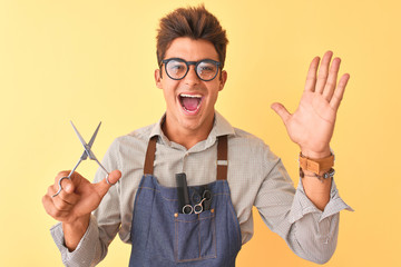 Handsome hairdresser man wearing apron holding scissors over isolated yellow background very happy and excited, winner expression celebrating victory screaming with big smile and raised hands